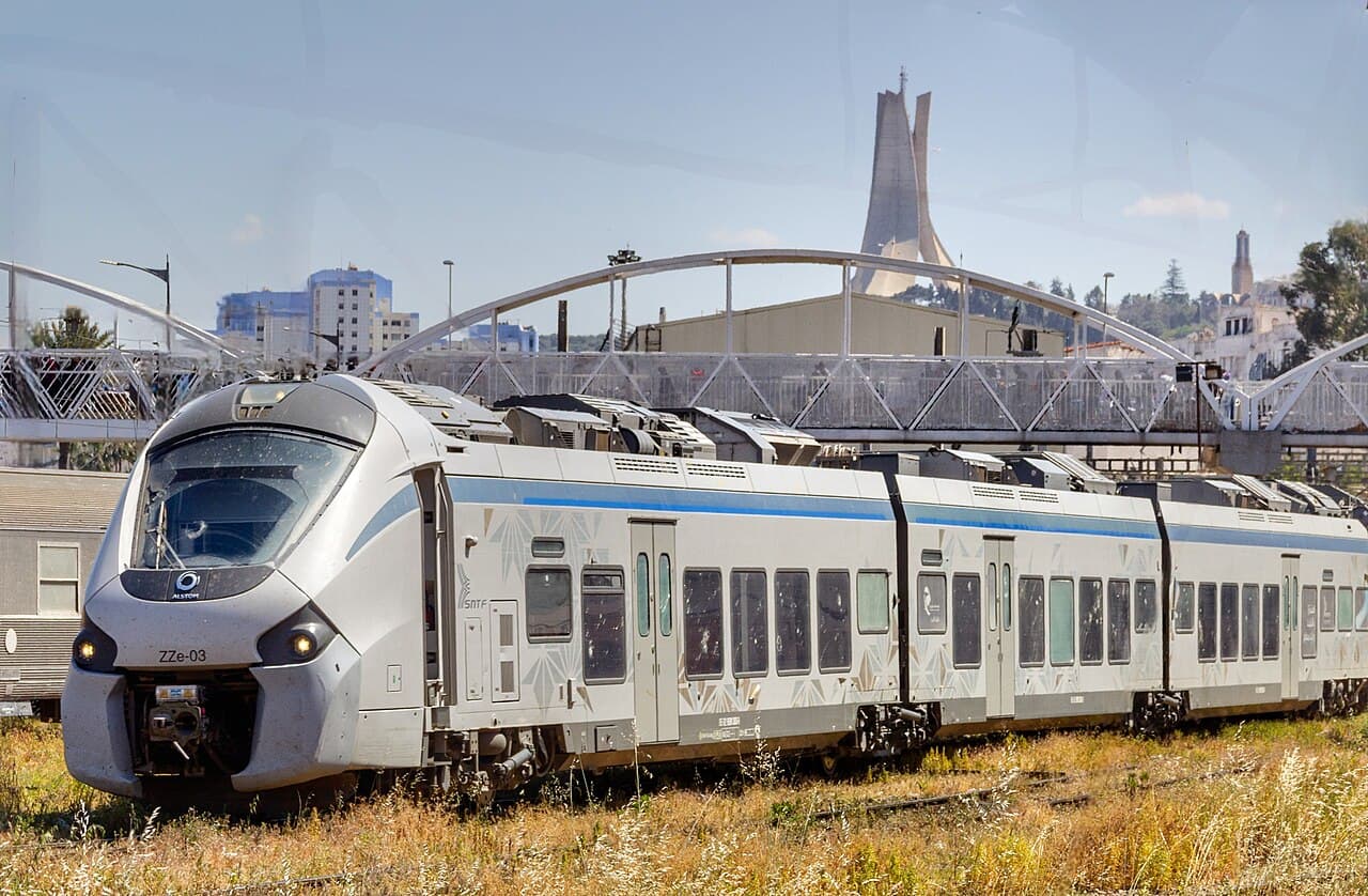 A modern passenger train at Agha station in Algiers, with Maqam Echahid on the skyline